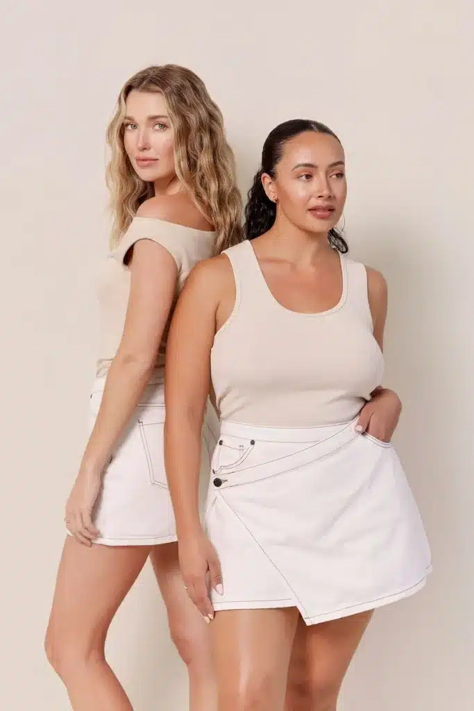 Two models posing back to back wearing neutral tank tops and off-white asymmetrical denim skorts