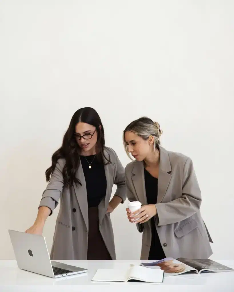 Two women wearing blazers standing at a desk and reviewing a laptop screen together